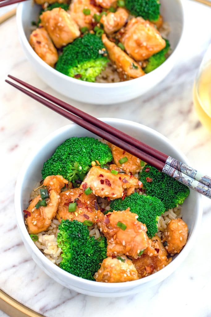 Overhead view of a white bowl of sesame chicken and broccoli over rice in a white bowl with chopsticks on a marble surface with a second bowl in the background