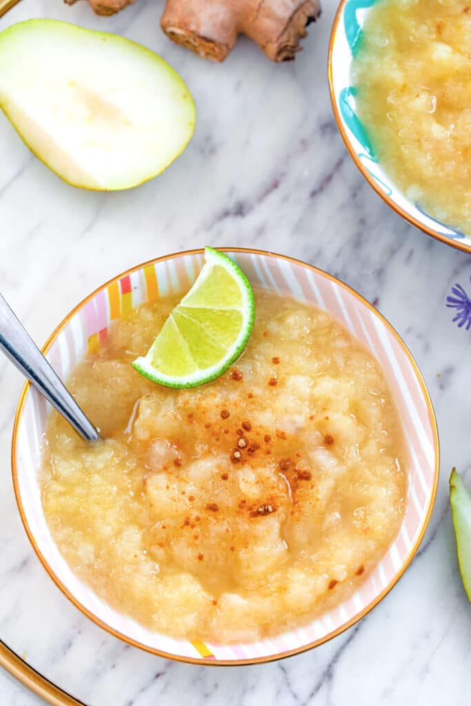 Overhead view of lime ginger pear sauce in a bowl topped with ground ginger and lime wedge with second bowl and pear half and fresh ginger in the background.