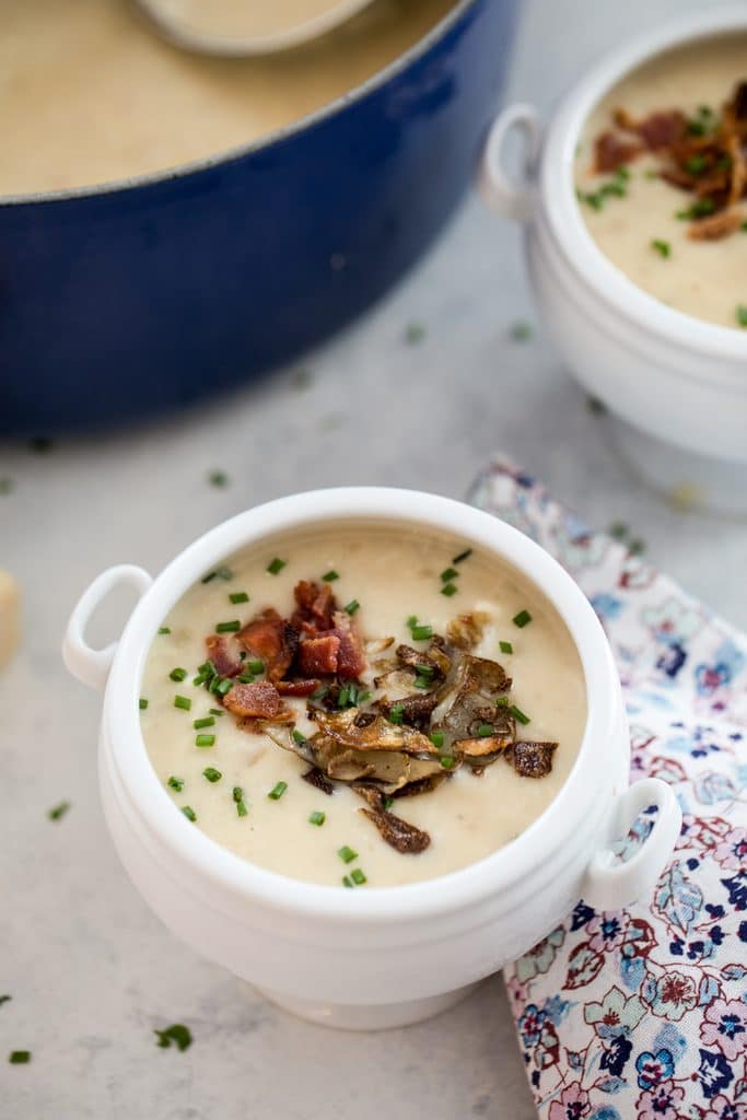 Overhead view of a white bowl of loaded baked potato soup with bacon, chives, and potato skins with second bowl or soup and pot of soup in background