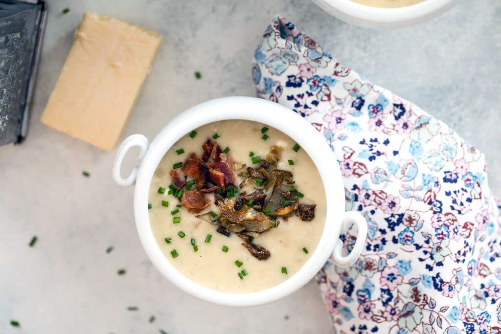 Landscape bird's eye view of a white bowl of loaded baked potato soup with crispy potato skins, bacon, and chives, with block of cheddar and cheese grater in background