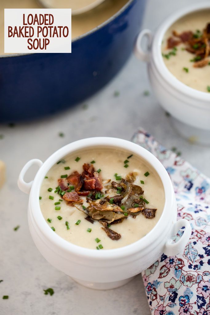 Overhead view of a white bowl of loaded baked potato soup with bacon, chives, and potato skins with second bowl or soup and pot of soup in background and recipe title at top