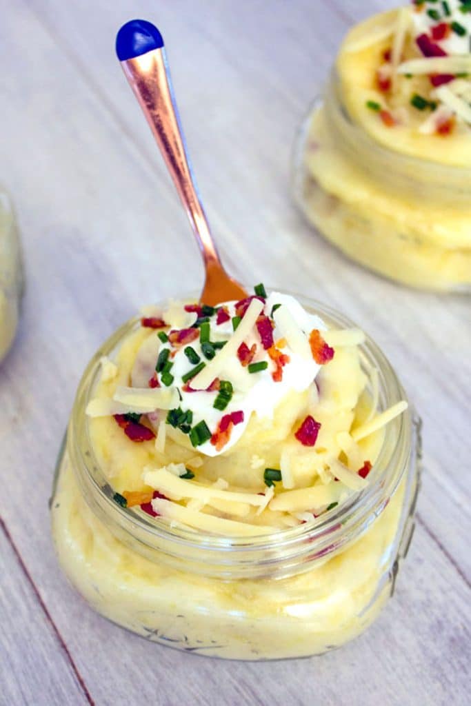 Overhead view of a small mason jar filled with mashed potatoes and topped with cheese, sour cream, chives, and crumbled bacon with a small gold fork sticking out of it and another jar in the background