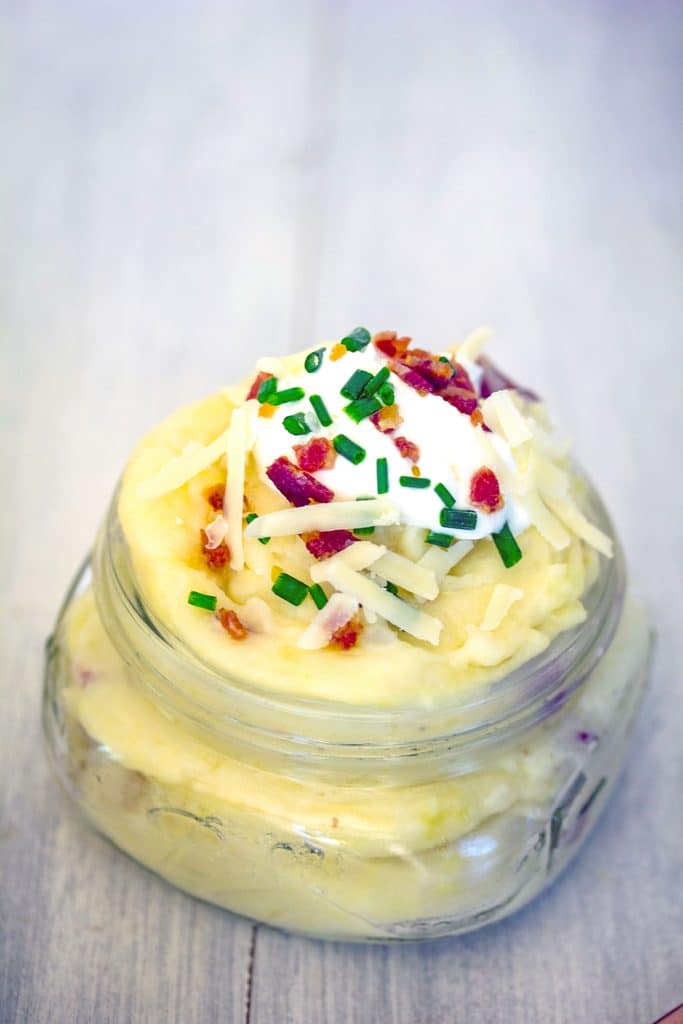 Close-up of one small mason jar of loaded mashed potatoes, topped with shredded cheese, sour cream, chives, and crumbled bacon