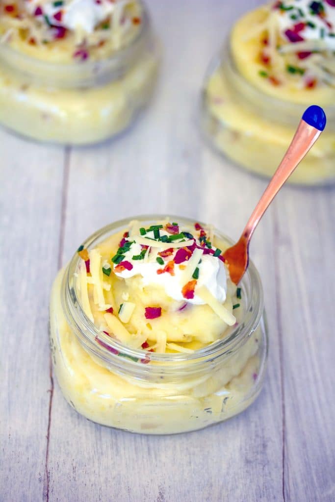 Overhead view of jar of loaded mashed potatoes with shredded cheese, sour cream, chives, and crumbled bacon with a small gold fork sticking out if it, with two more loaded mashed potato jars in the background