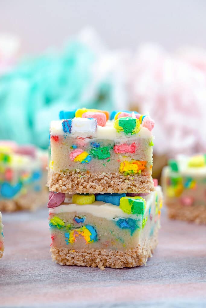 Head-on closeup view of two Lucky Charms cookie dough bars with cereal crust and buttercream frosting with more bars in the background