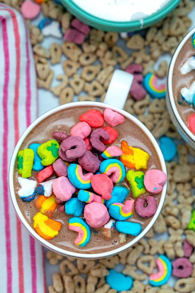 Overhead view of mug of Lucky Charms hot chocolate topped with cereal marshmallows with cereal all around.
