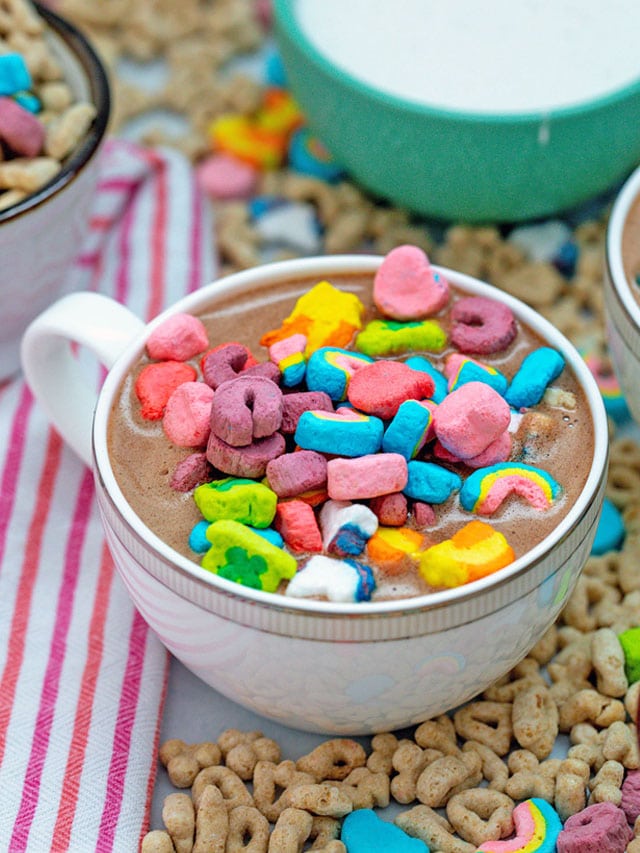 Lucky Charms Hot Chocolate in a coffee cup with cereal all around and marshmallow fluff in background.
