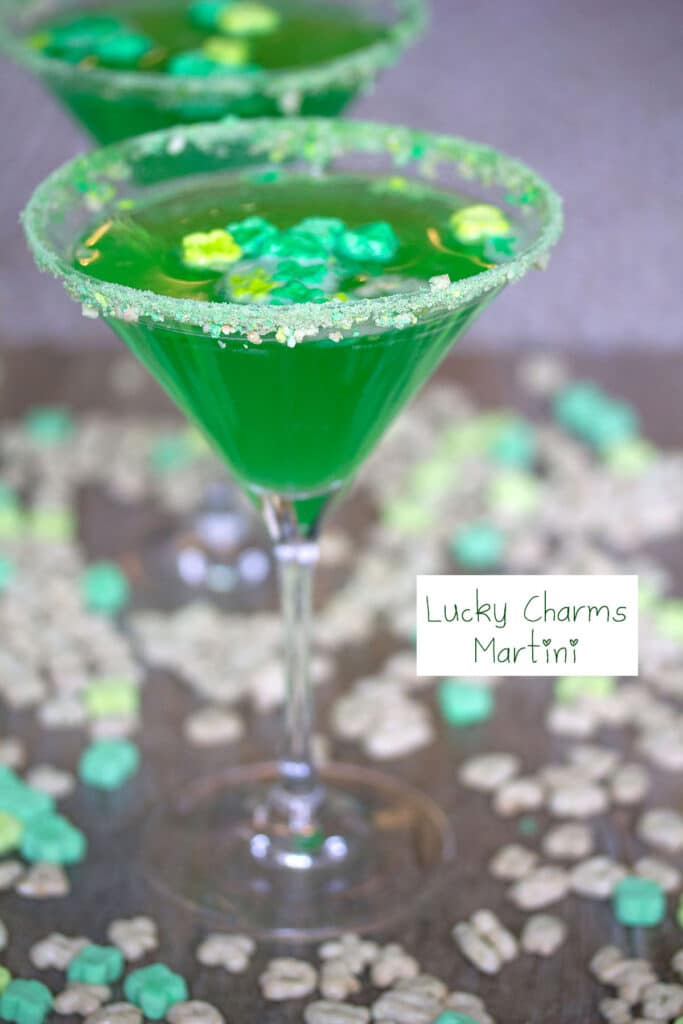 Head-on view of green Lucky Charms Martini with Lucky Charms cereal and second martini in background and recipe title on side