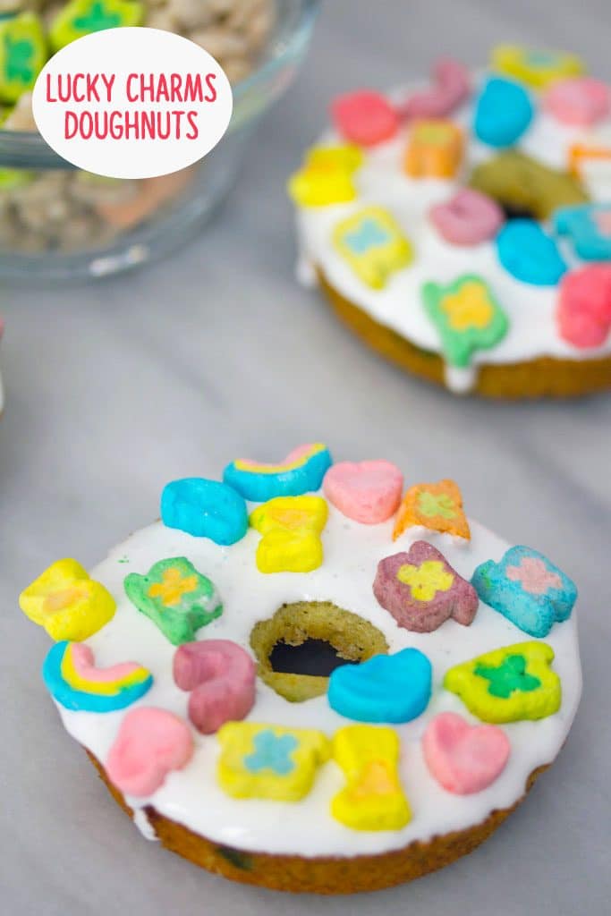 Overhead view of Lucky Charms doughnuts on a marble surface with bowl of Lucky Charms in the background and recipe title at top