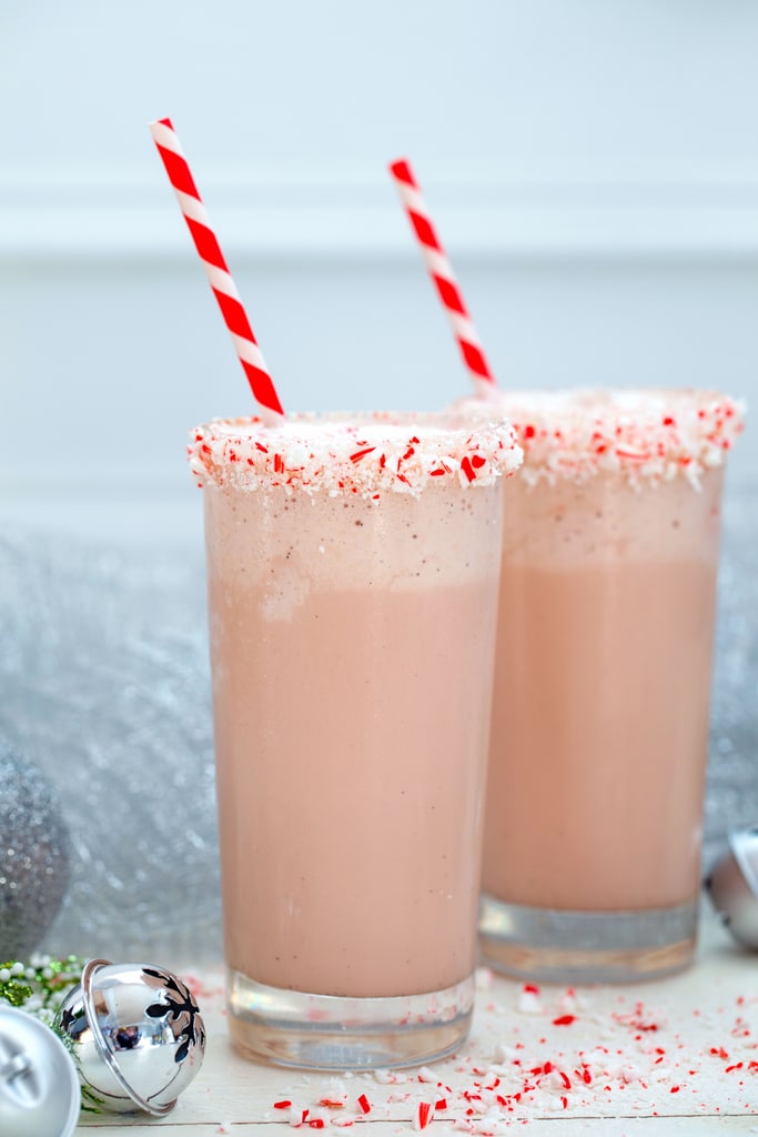 Head-on closeup of two malted candy cane milkshakes with crushed candy cane rims and red and white straws with silver bell and crushed candy canes all around