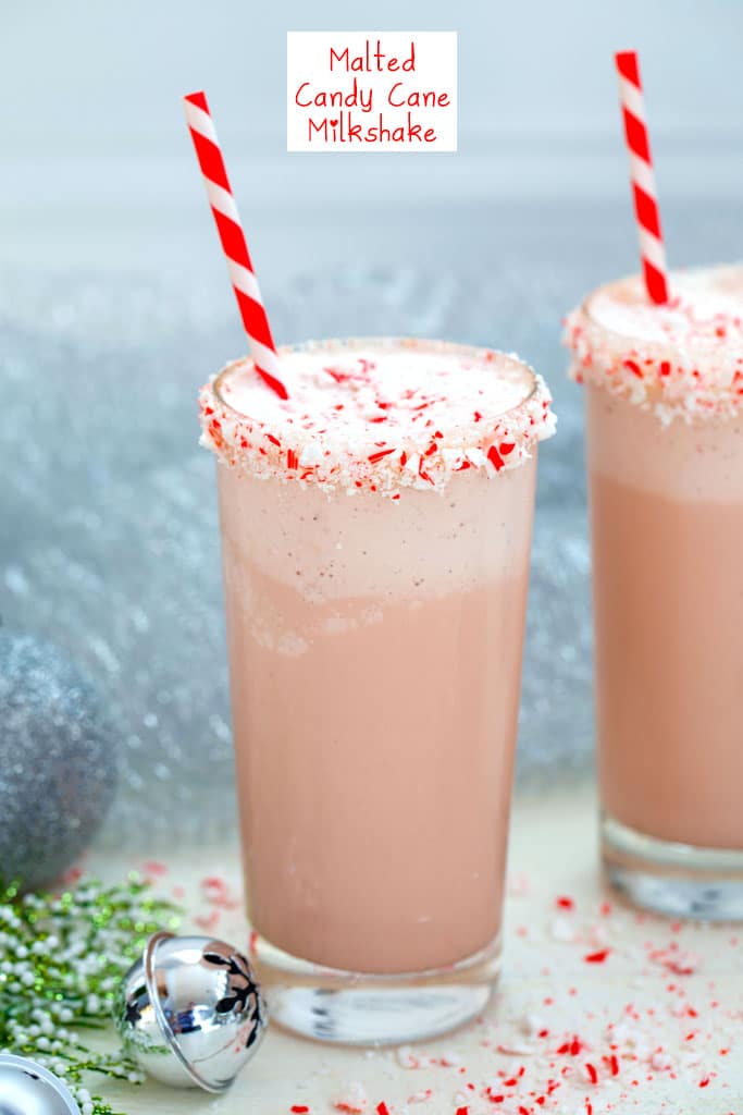 Head-on view of a malted candy cane milkshake with crushed candy cane rim with red and white striped straw with second milkshake, silver bells, and holly in background with recipe title at top