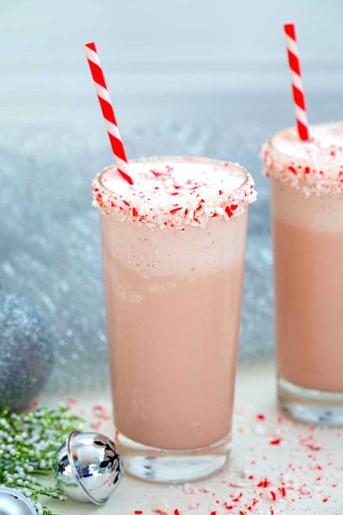 Head-on view of a malted candy cane milkshake with crushed candy cane rim with red and white striped straw with second milkshake, silver bells, and holly in background