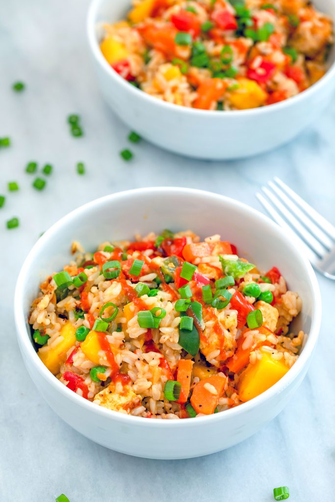 Head-on view of a bowl of mango coconut fried rice with second bowl of rice, scallions, and fork in the background