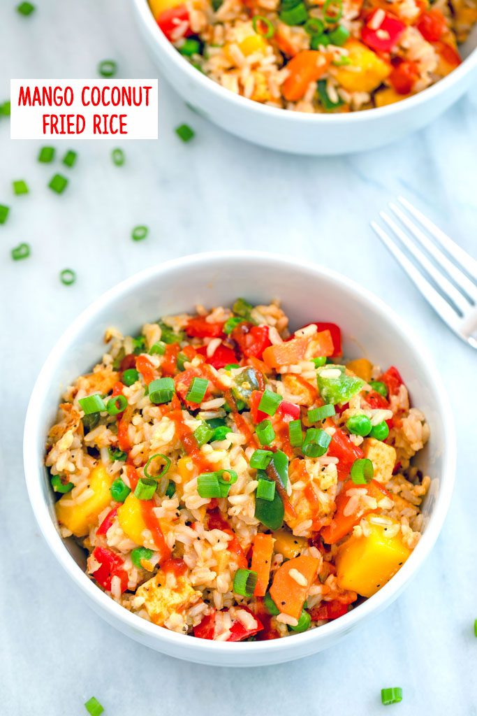 Overhead view of a bowl of mango coconut fried rice on marble surface with second bowl, chopped scallions, and fork in the background with recipe title at top