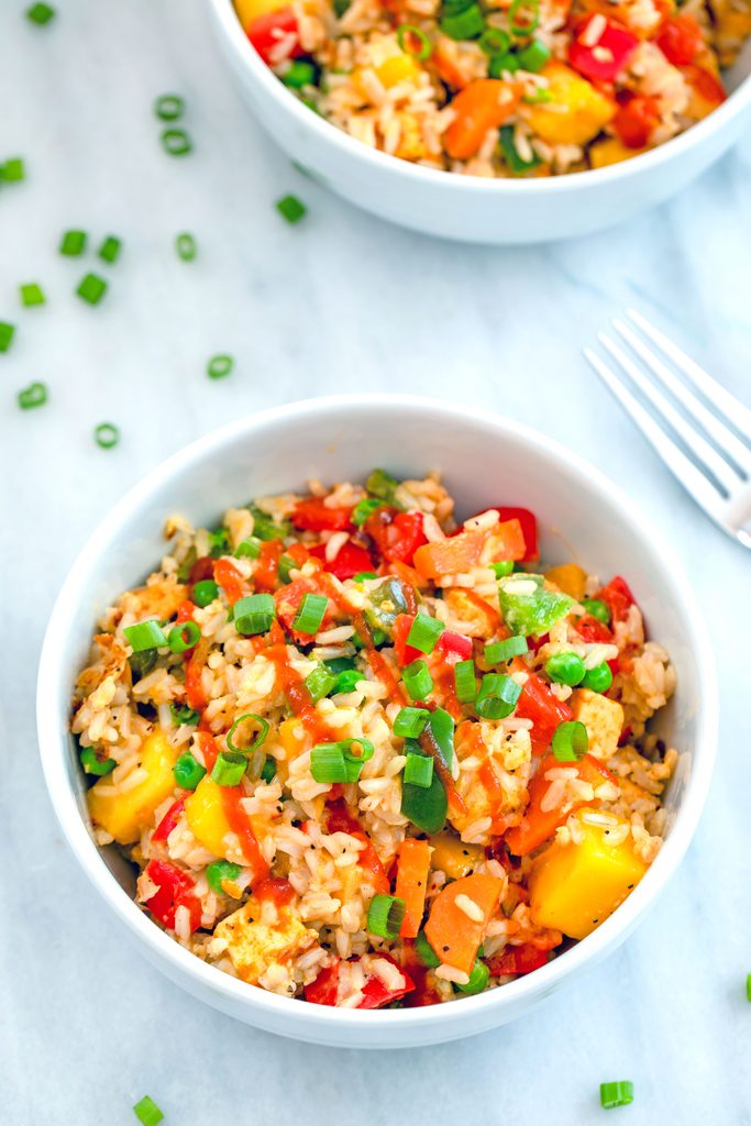 Overhead view of a bowl of mango coconut fried rice on marble surface with second bowl, chopped scallions, and fork in the background