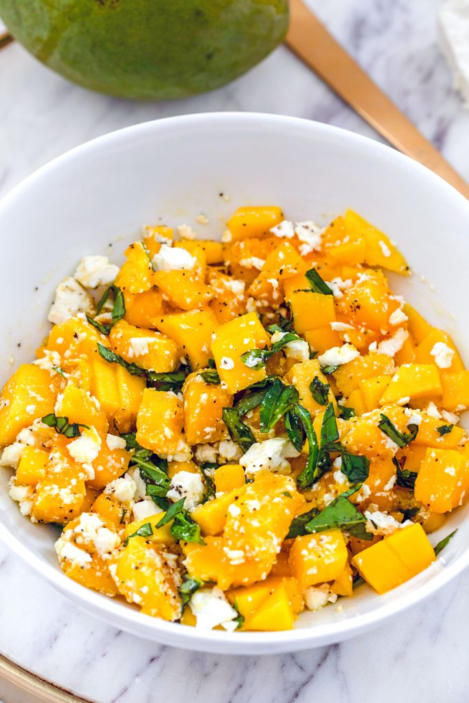 Head-on view of a white bowl with mango and feta salad with basil on a marble surface with mango and gold serving piece in the background