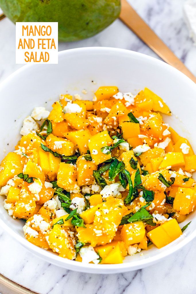 Head-on view of a white bowl with mango and feta salad with basil on a marble surface with mango and gold serving piece in the background and recipe title at top