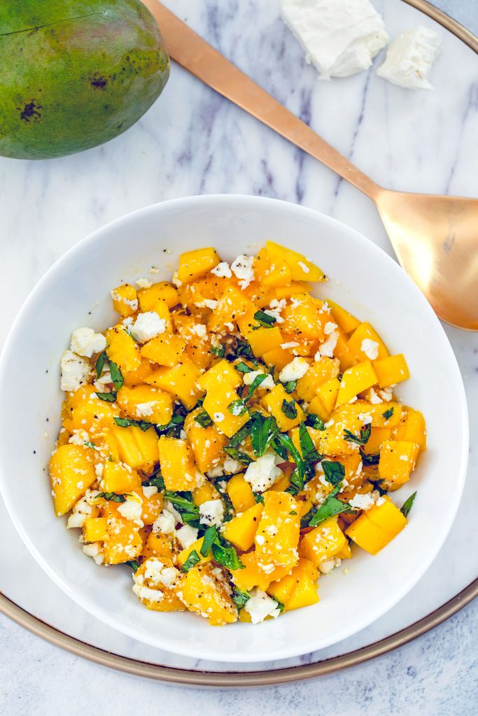 Overhead view of a white bowl of mango and feta salad with basil on a marble tray with gold serving spoon, mango, and feta cheese in the background