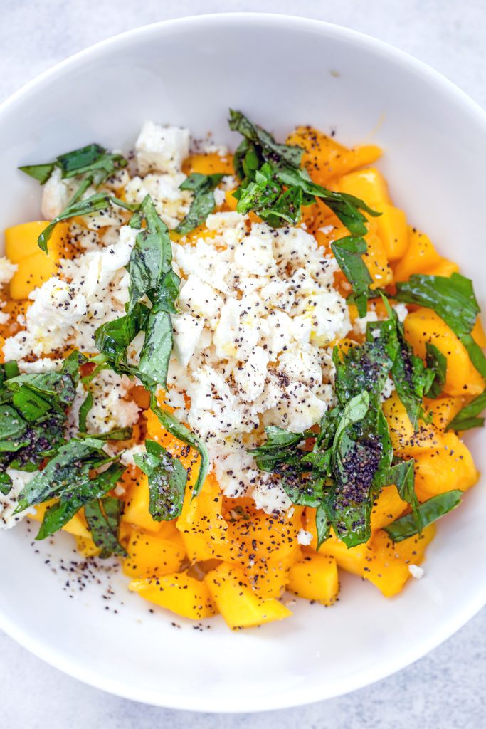 Overhead view of ingredients for mango and feta salad in a bowl, including chopped mango, sliced basil, feta, olive oil, and salt and pepper