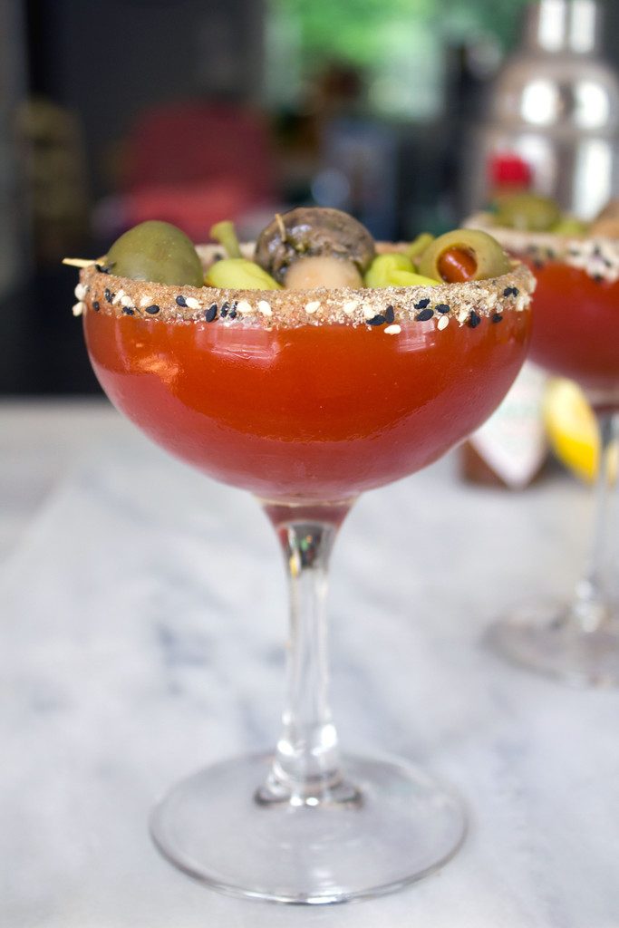 Head-on closeup view of a dirty bloody mary martini with second martini and cocktail shaker in background