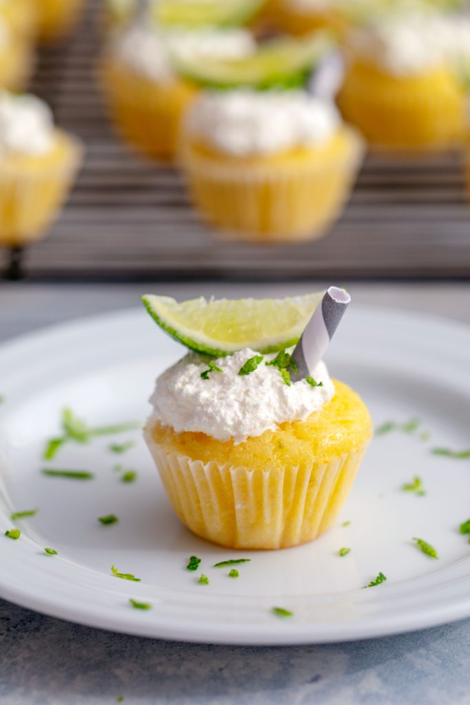 Head-on view of a mini margarita cupcake on a white plate with lime zest, lime wedge, and mini straw with more cupcakes in background and recipe title at top