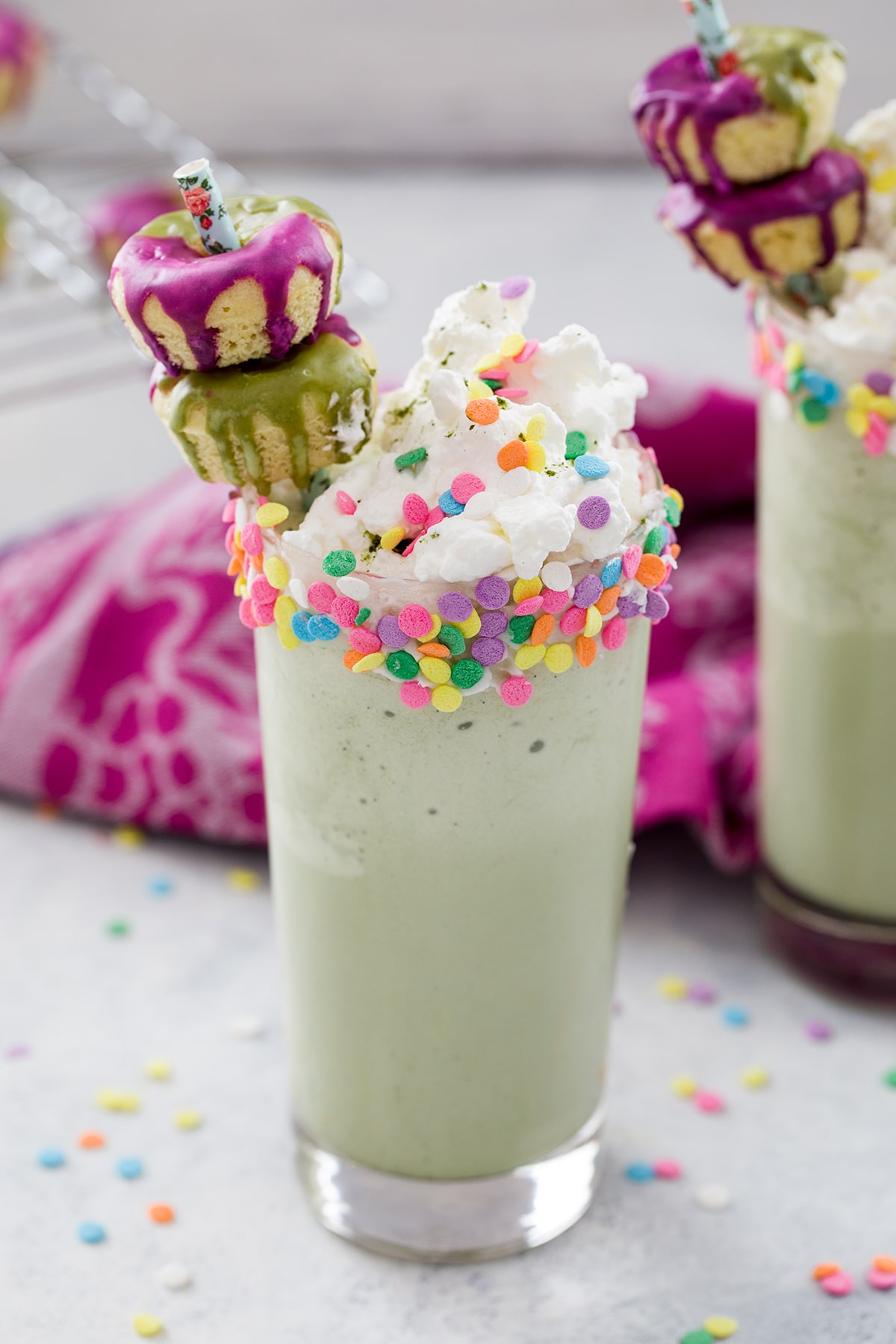 Overhead view of matcha milkshake with sprinkles rim, whipped cream, and mini donut garnish with second milkshake in the background and sprinkles all around.