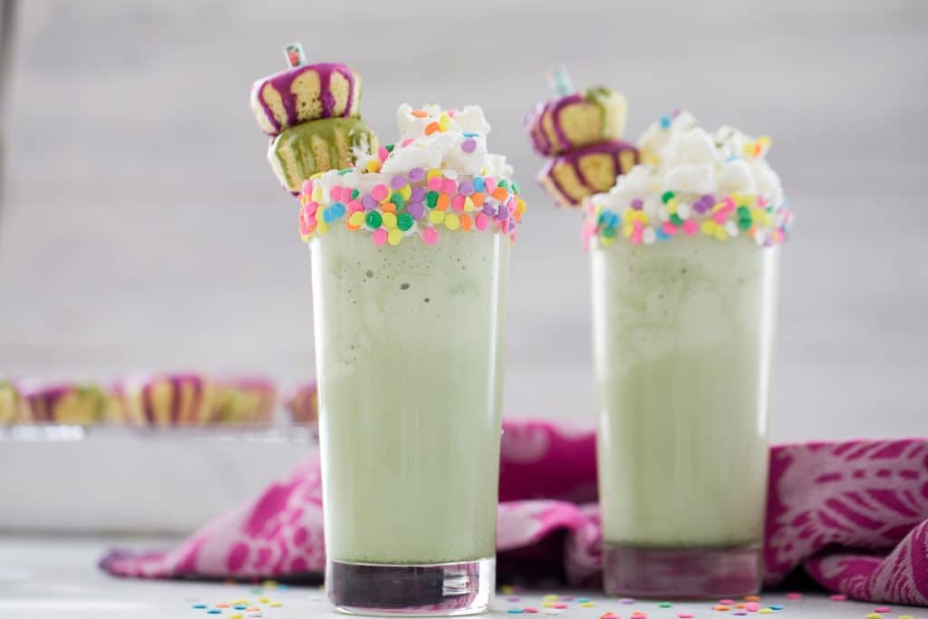 Landscape head-on view of two matcha milkshakes with sprinkles on the rim, whipped cream, and donut garnishes with more donuts on rack in the background