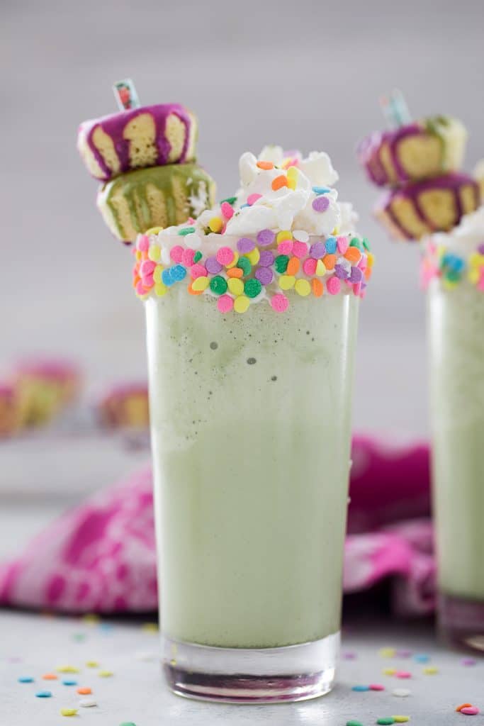 Head-on view of a matcha milkshake with sprinkles on the rim, whipped cream, and mini donut garnish with second milkshake, sprinkles, and donuts in the background