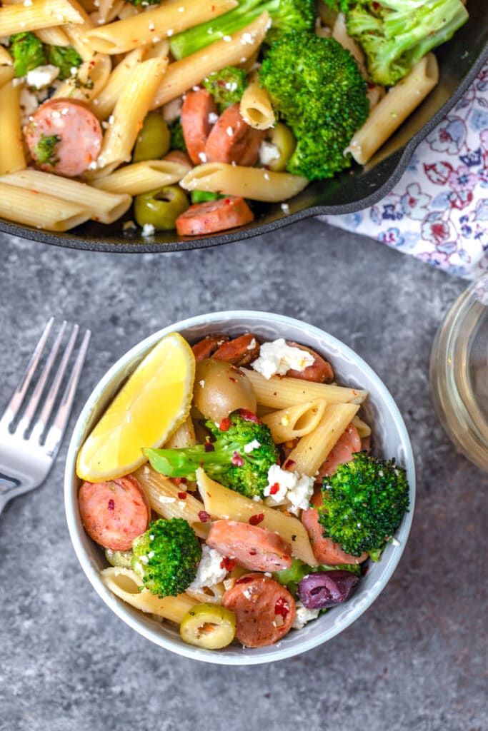 Overhead view of bowl of Mediterranean pasta with chicken sausage, broccoli, olives, and feta with fork and skillet in background