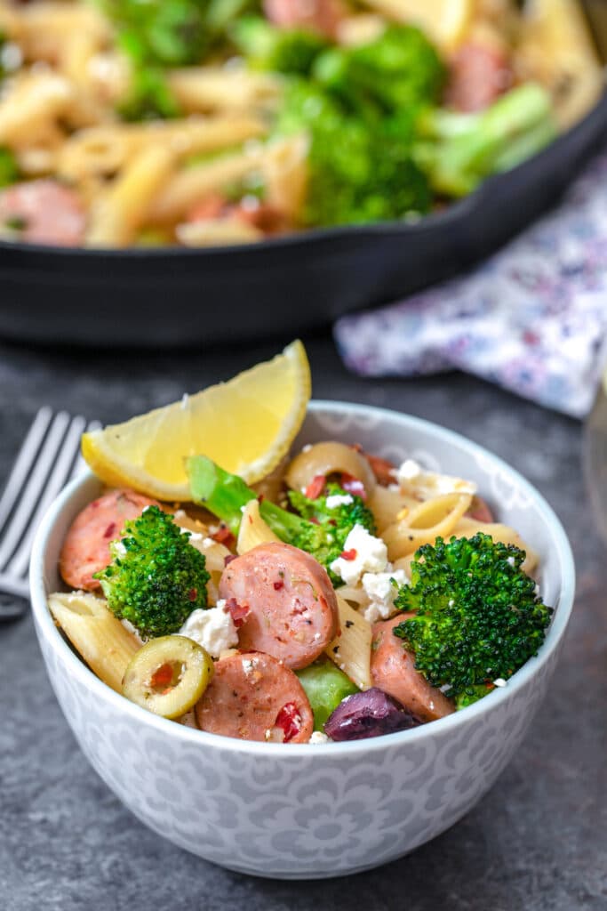 Head-on closeup view of a bowl of Mediterranean pasta with chicken sausage, broccoli, olives, feta, and lemon with skillet in background