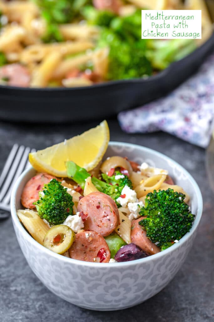 Head-on closeup view of a bowl of Mediterranean pasta with chicken sausage, broccoli, olives, feta, and lemon with skillet in background and recipe title at top