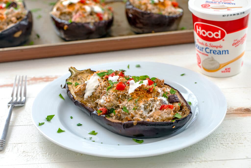 Landscape view of Mediterranean turkey stuffed eggplant half on a white plate with fork, container of Hood Sour Cream, and more eggplants on a baking sheet in the background