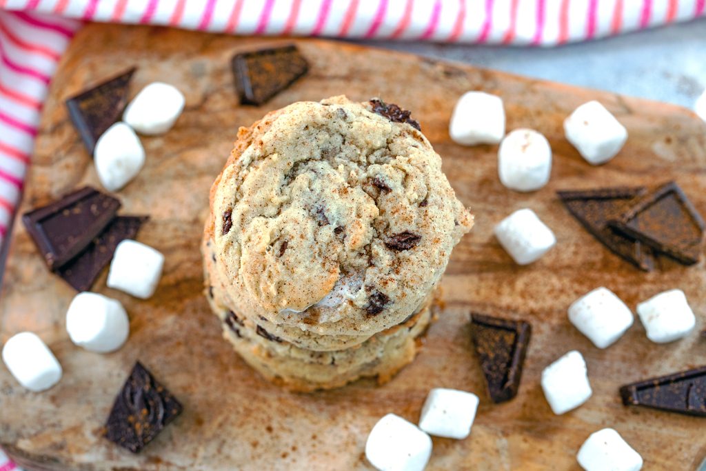 Landscape view of Mexican chocolate cookies stacked on top of each other on a wooden board with dark chocolate chunks and mini marshmallows all around and cinnamon sprinkled on top with red and pink striped towel in background