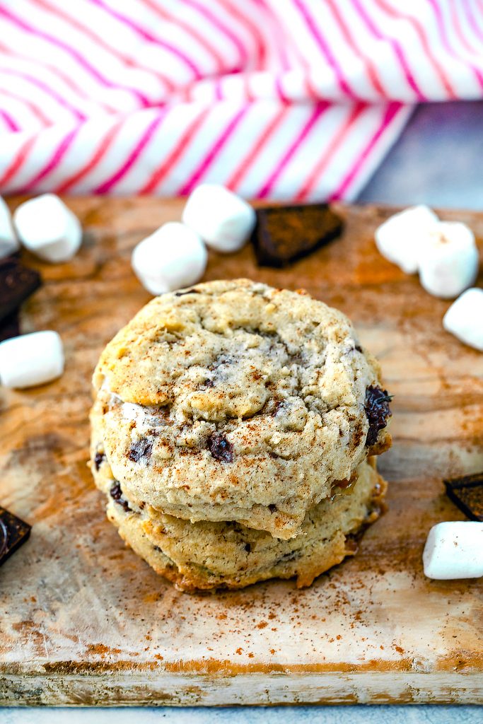 Head-on view of three Mexican chocolate cookies stacked on top of each other on wooden board with mini marshmallows and chopped dark chocolate scattered around and cinnamon sprinkled on top