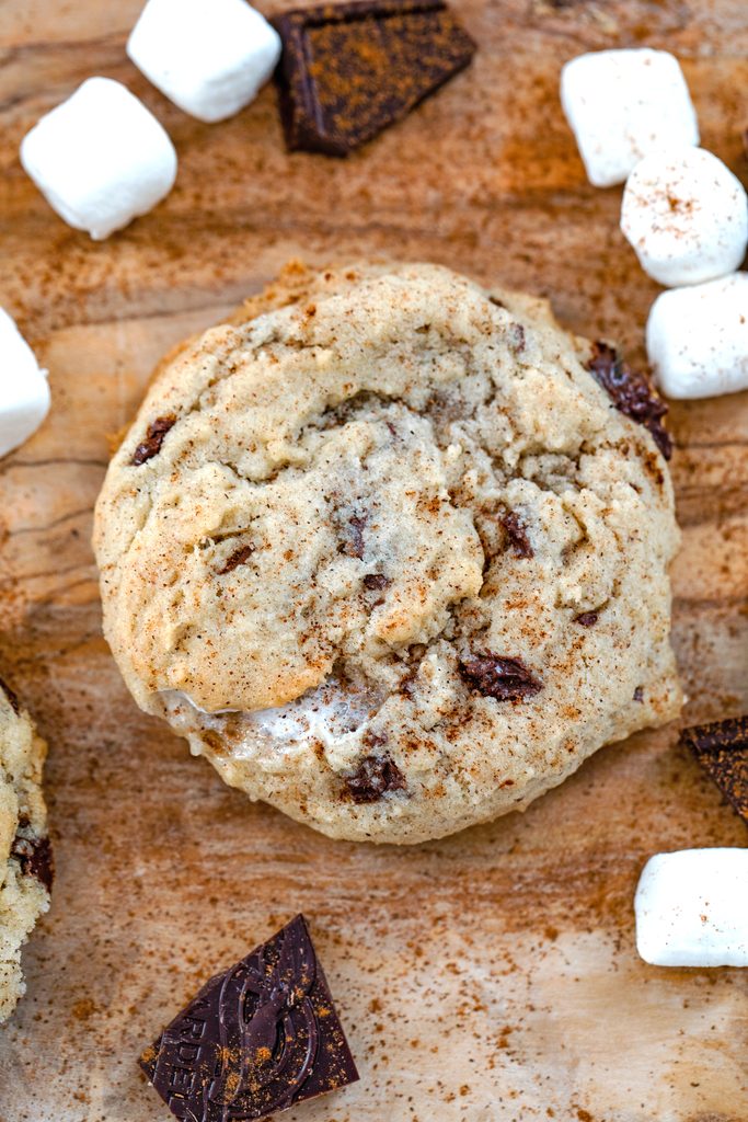 Overhead view of a Mexican chocolate cookie on a wooden surface with mini marshmallows and dark chocolate chunks all around and cinnamon sprinkled on top