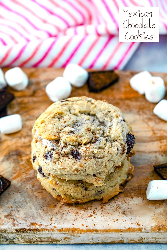 Head-on view of three Mexican chocolate cookies stacked on top of each other on wooden board with mini marshmallows and chopped dark chocolate scattered around and cinnamon sprinkled on top with recipe title at top of image