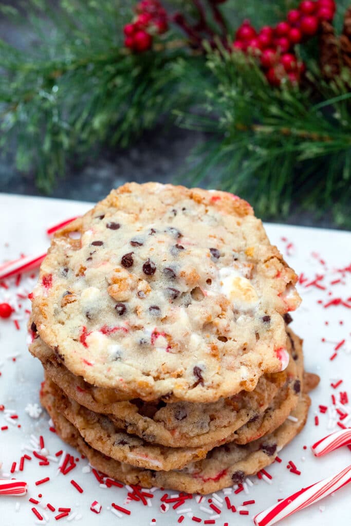 View of a stack of Milk Bar's Cornflake Chocolate Chip Candy Cane Cookies with sprinkles and candy canes all around and holly in background