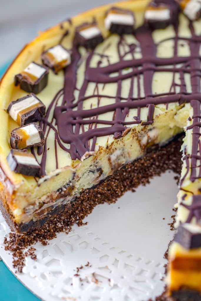 Close-up of entire Milky Way cheesecake with chocolate drizzle with a slice taken out on a white doily on a turquoise cake stand