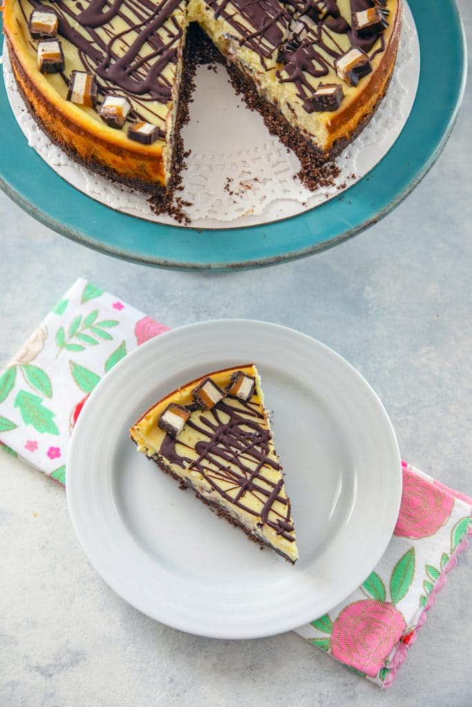 Bird's eye view of slice of Milky Way cheesecake on a white plate and pink flowered towel with rest of cheesecake on a turquoise cake stand in the background