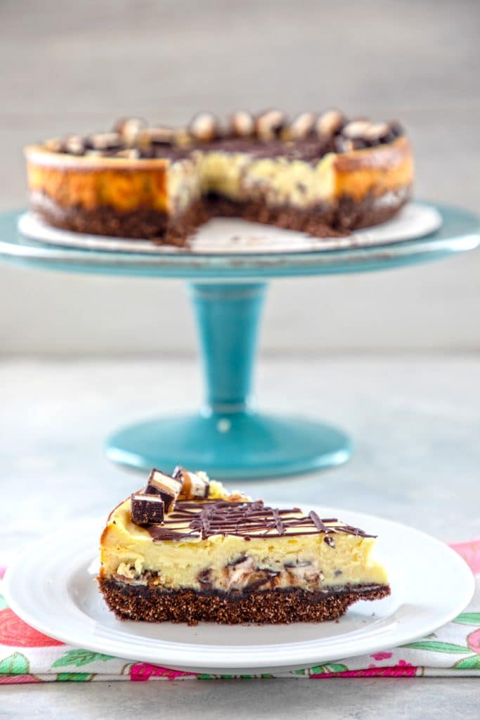 Head- on view of milky way cheesecake with a slice on a white plate and the rest of the cheesecake on an aqua cake stand in the background