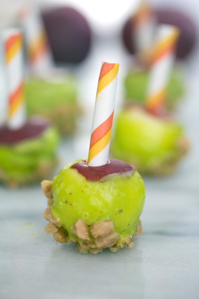 Head-on view of a mini caramel candy apple coated in green candy melts with heath bar crunch with more mini candy apples in the background