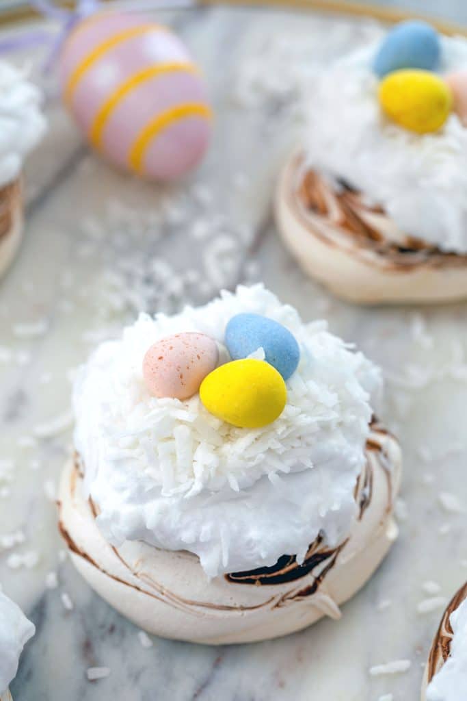 Head-on closeup of a chocolate swirled mini Easter pavlova topped with whipped coconut cream, shredded coconut, and Cadbury Mini Eggs on marble surface with Easter eggs and more pavlova in background
