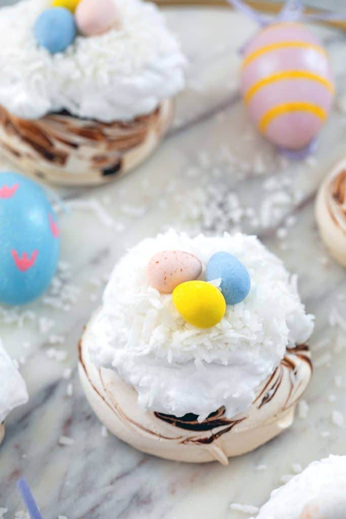 Head-on view of chocolate swirled mini Easter pavlovas topped with whipped cream, coconut, and Cadbury Mini Eggs on a marble surface with Easter eggs in background
