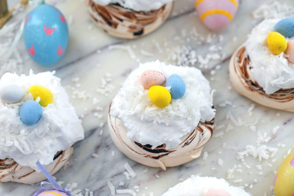 Landscape overhead view of multiple chocolate swirled mini Easter pavlovas topped with coconut whipped cream, shredded coconut, and Cadbury Mini Eggs on a marble surface surrounded by painted Easter eggs