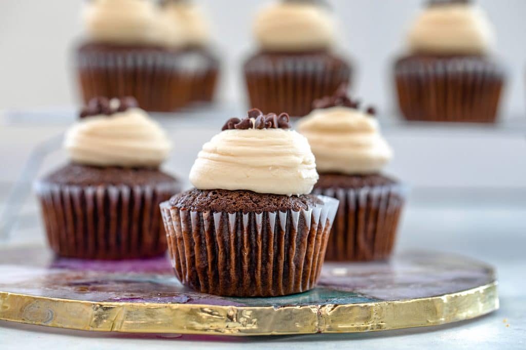 Landscape view of chocolate mint chocolate chip cupcakes topped with Baileys frosting and mini chocolate chips on a gold rimmed platter with more cupcakes in the background