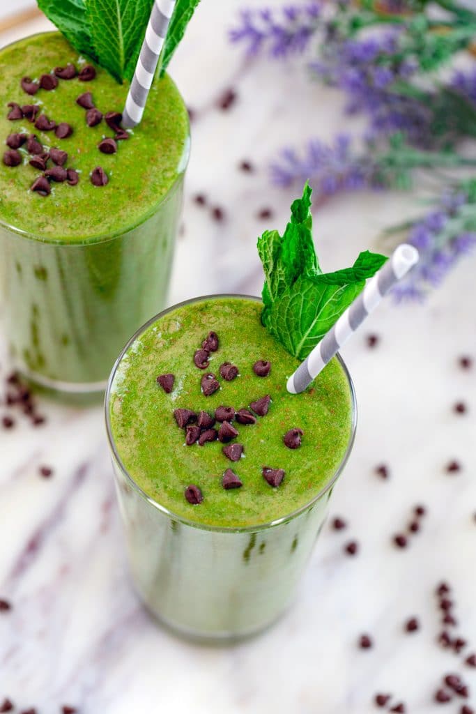 Overhead view of two mint chocolate green smoothies topped with mini chocolate chips and mint garnish on a marble surface with mini chocolate chips and purple flowers