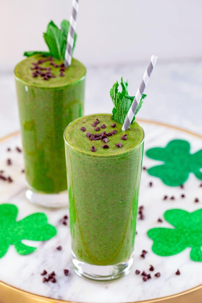 Head-on view of two mint chocolate green smoothies on a marble platter topped with mini chocolate chips and mint garnish with glitter shamrocks and mini chocolate chips in background