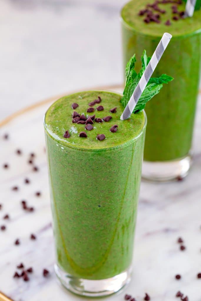 Head-on view of mint chocolate green smoothie topped with mini chocolate chips and mint garnish on a marble platter with a second smoothie in the background and recipe title at top