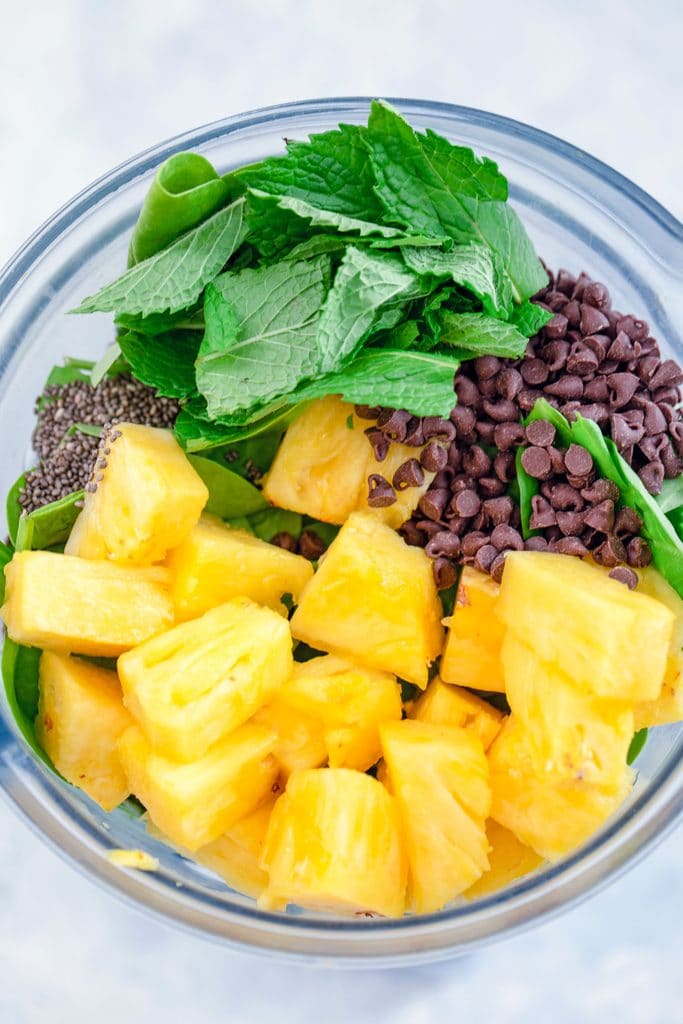 Overhead view of milk, spinach, pineapple, mini chocolate chips, mint, and chia seeds in a blender