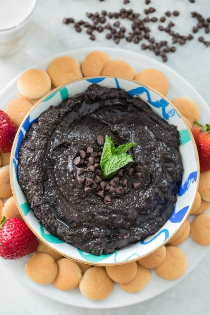 Close-up bird's eye view of bowl of mint chocolate hummus topped with mini chocolate chips and mint leaves on a white plate with mini Nilla Wafers and strawberries and mini chocolate chips in the background